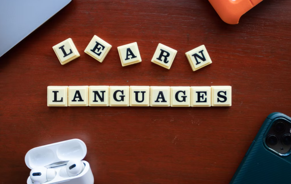 This image shows Scrabblk=a mobile, hands free, laptop and word ‘learn languages’ have kept on brown table